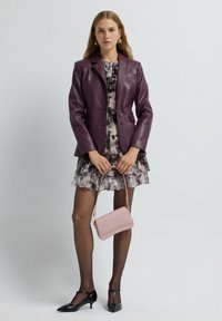 Blazer viola in ecopelle sopra un vestito ricamato con volant. Borsa rosa con accenti dorati; décolleté neri con tacchi kitten. Texture lisce e design aderente.