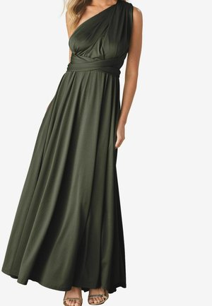 Femme portant une robe longue vert foncé à une épaule avec une taille enveloppée, assortie à des sandales dorées à bouts ouverts.