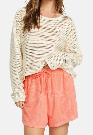 Kvinde iført en cremefarvet åbenstrikket sweater og koralfarvede shorts med snøre og et subtilt palmebladsmønster, stående mod en ensfarvet baggrund.
