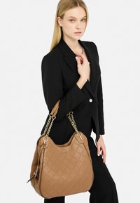 Sac à main en cuir marron avec un motif matelassé et des anses en chaîne dorée, arborant un logo doré et une silhouette courbée.