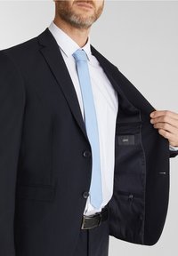 Blazer blu navy con due bottoni, indossato sopra una camicia bianca con pois e una cravatta blu chiaro. L'interno ha una tasca con un'etichetta del marchio visibile.