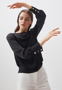 Blusa nera a maniche lunghe con una vestibilità ampia; presenta dettagli con bottoni dorati sui polsini; texture liscia e design fluido.