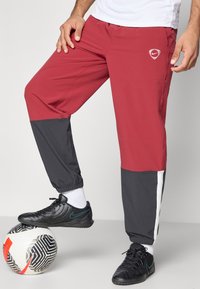 Κόκκινες και γκρίζες αθλητικές παντελόνες με ελαστικές μανσέτες, που διαθέτουν λευκό λογότυπο στο μηρό, συνδυασμένες με μαύρες ποδοσφαιρικές παπούτσια και μια μπάλα ποδοσφαίρου.