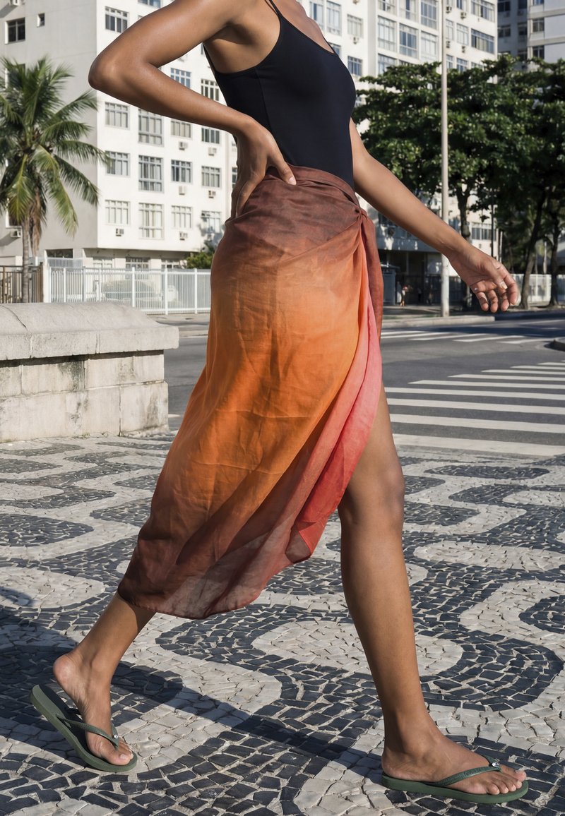 Persona che cammina su un pavimento a motivo indossando un costume da bagno nero, gonna a portafoglio arancione-rossa e infradito verdi in un ambiente urbano esterno.
