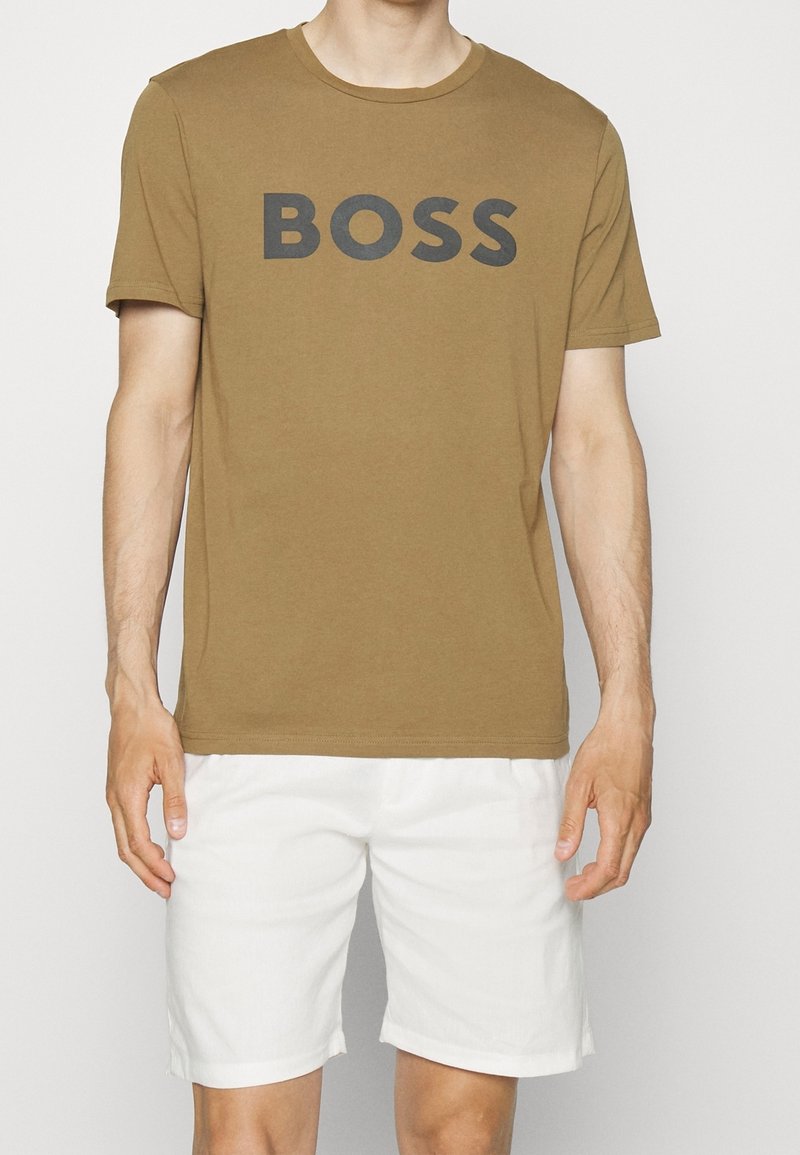 Homme portant un T-shirt marron avec le mot BOSS en grandes lettres et un short blanc jusqu'aux genoux.
