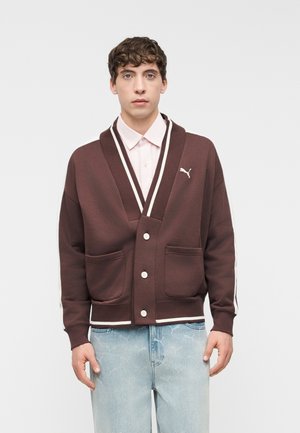 Νέος άνδρας που φοράει ένα καφέ ζακέτα Puma με λευκή περίγραμμα και ανοιχτά μπλε τζιν, στέκεται μπροστά σε ένα απλό φωτεινό φόντο.