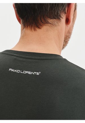 Ciemnozielona koszulka z okrągłym dekoltem i krótkimi rękawami, z białym logo "PAKO LORENTE" nadrukowanym na górnej plecach.