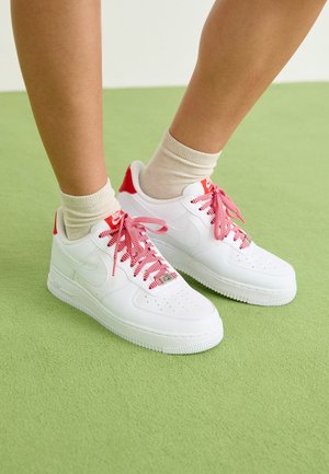 White leather sneakers with red accents and laces, featuring a perforated toe box and a rubber sole. Socks are beige and ribbed.