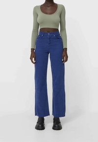 Blaue Cord-Röhrenjeans mit weitem Bein, hoher Taille, Fronttaschen und rohem Saum; kombiniert mit einem figurbetonten, langärmeligen, olivgrünen Oberteil.