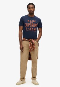 Camiseta de color navy con texto gráfico naranja, pantalones de pana beige y una camisa de cuadros atada a la cintura. Zapatillas blancas completan el conjunto.