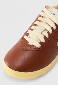 Brązowy skórzany sneaker z fakturowanym wykończeniem, beżowymi sznurowadłami i kremową gumową podeszwą. Charakteryzuje się przeszywanym detalem w okolicy palców.