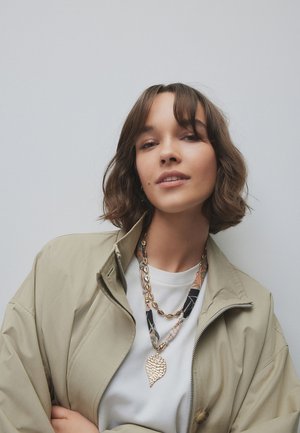 Junge Frau mit kurzen braunen Haaren trägt eine beige Jacke über einem weißen Hemd und mehreren geschichteten Halsketten, posiert vor einem einfarbigen hellgrauen Hintergrund.