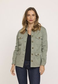 Veste utilitaire vert olive avec boutons sur le devant, quatre poches et manches longues. Fabriquée en coton, elle présente une texture lisse et un design léger.