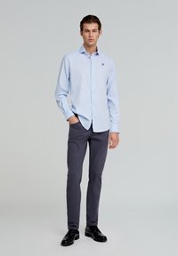 Himmelblaues Langarmhemd mit Button-Down-Kragen, tonalem Logo auf der Brust, kombiniert mit grauen Slim-Fit-Hosen und schwarzen Pumps.