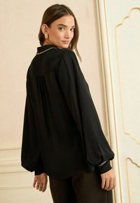 Černá halenka s dlouhým rukávem a volným střihem, s nařasením na zadní straně a ozdobnými manžetami. Hladká látka s jemným leskem.