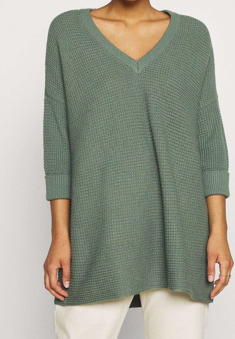 Femme portant un pull ample en maille verte avec col en V et manches trois-quarts, associé à un pantalon de couleur claire.