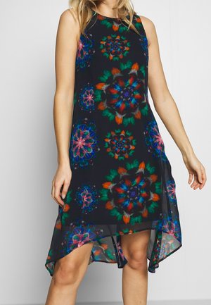 Hihaton musta mekko, jossa värikkäät symmetriset kukkakuviot ja etuosa lyhyempi kuin takaosa; mekkoa pitää yllään seisova henkilö, jonka kädet ovat rentoina sivuilla.