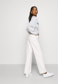 Sweatshirt oversized cinza claro com gráfico azul, combinado com calças largas em tom creme e ténis brancos numa pose de perfil contra um fundo simples.