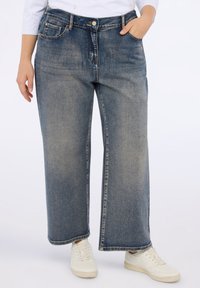 Jeans de denim de pierna ancha en un lavado azul desgastado. Presentan una cintura alta, un diseño tradicional de cinco bolsillos y costuras visibles. Combinados con zapatillas blancas.
