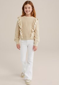 Beige stickad tröja med volangdetaljer, kombinerad med vita utsvängda jeans och ljusa sneakers. Enkel design och texturerad tyg.