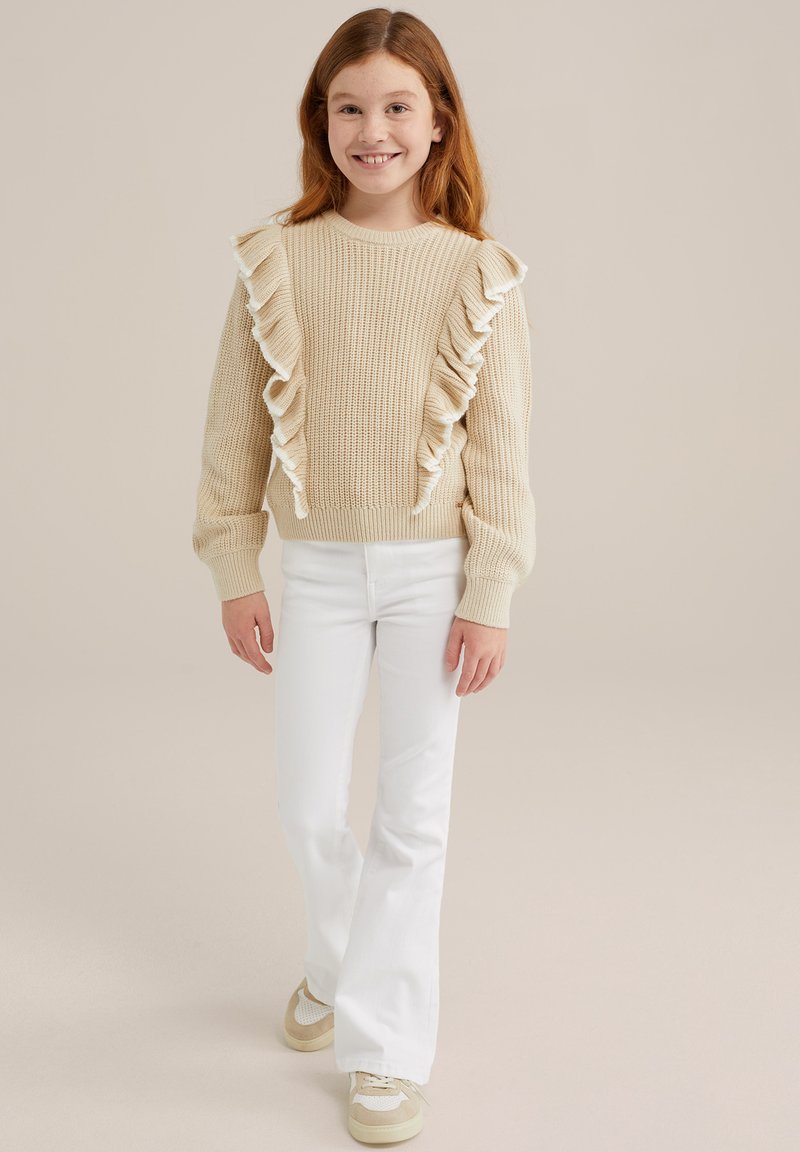 Beige stickad tröja med volangdetaljer, kombinerad med vita utsvängda jeans och ljusa sneakers. Enkel design och texturerad tyg.