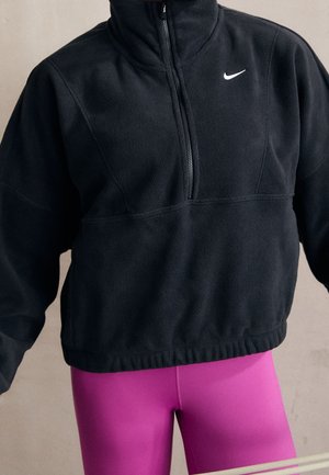 Persona con un forro polar negro de Nike con cremallera hasta un cuarto y mallas de color rosa brillante, con los brazos ligeramente levantados, mostrada desde la barbilla hasta mitad del muslo.