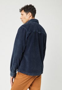 Camisa de pana azul marino con cuello, mangas largas y puños abotonados. Presenta detalles en el yugo trasero y un dobladillo sin acabar.