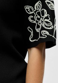 Haut en maille noir à manches courtes avec broderie florale blanche sur la manche, montrant une partie d'un bras nu et des détails de tissu texturé.