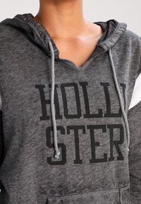 Šedá mikina s výstřihem do V, na přední straně velký nápis "HOLLISTER" černým písmem. Obsahuje stahovací šňůrky a texturovanou látku s bílými detaily.