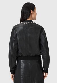 Giubbotto bomber in pelle nera con vita aderente e schiena decorata con borchie metalliche lungo il colletto. Texture liscia, design casual.