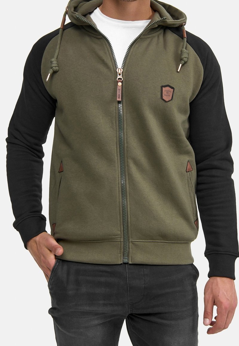 Homme portant un sweat à capuche zippé vert olive avec manches noires, cordons et petit emblème sur la poitrine, associé à un jean foncé et un sous-vêtement blanc.