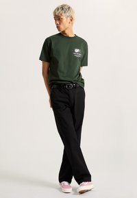 Camiseta de algodón verde con un estampado gráfico blanco en el lado izquierdo del pecho, combinada con pantalones rectos negros y zapatillas rosas.
