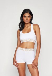 Belo atletski komplet, ki vključuje športni modrček z okrolim izrezom in hlačke v slogu boyshorts, oba z zlatim grškim ključnim vzorcem in logotipom na pasu. Gladek tekstura.