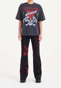 Černé riflové kalhoty s rozšířenými nohavicemi a červenou grafikou, včetně srdcí a lebek. V kombinaci s tmavým tričkem s grafikou, na kterém je lebka a živé vzory.