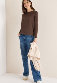 Bruine blouse met lange mouwen, gecombineerd met lichtblauwe losse jeans. De persoon houdt een beige jas vast en draagt witte sneakers met een beige zool.