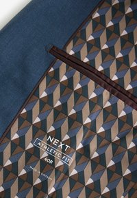 Granatowy materiał z wzorzystym wnętrzem, na którym znajdują się geometryczne kształty w odcieniach brązu, zieleni i niebieskiego. Metka informuje "NEXT ATHLETIC FIT 40R."