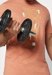Zwarte dumbbell vastgehouden in een hand; draagt een licht perzikkleurig T-shirt met "JUST DO IT" in geel en wit. Bevat een Nike-logo onderaan links.