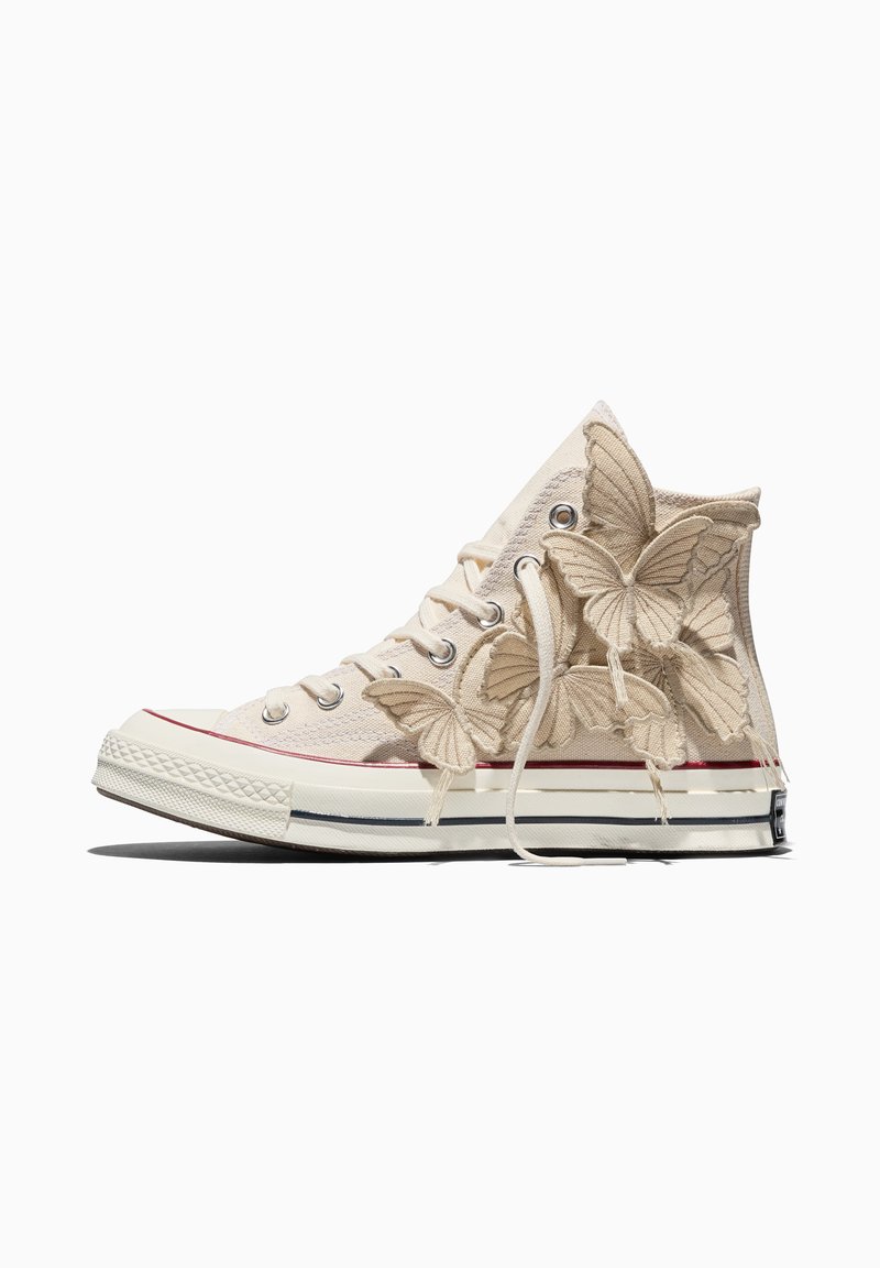 Beiger High-Top-Sneaker mit Stoff-Schmetterlingsapplikationen an der Seite und weißen Schnürsenkeln, liegend auf einer weißen Oberfläche.