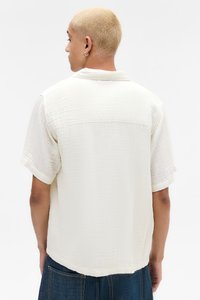 Kortermet, kremfarget skjorte med en teksturert, rynket finish; har krage og rett nederkant. Brukt med mørk denimshorts.