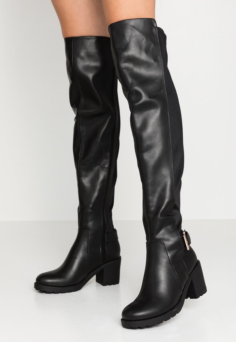Bottes hautes en cuir noir au-dessus du genou avec talon bloc, texture lisse, détail zippé et une sangle d'accent avec ferrures sur le côté.