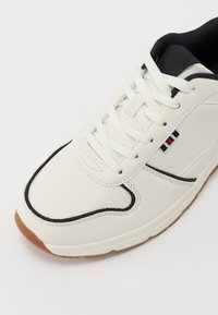 White leather sneaker with black accents and textured panels. Features white laces and a rubber gum sole. Side branding detail included.