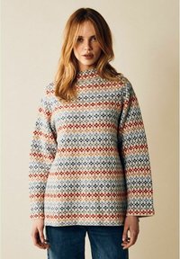 Kvinna med ljusbrunt hår som bär en långärmad stickad tröja med mönstrade ränder i röd, blå och beige samt blå jeans.