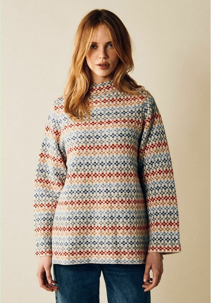 Kvinna med ljusbrunt hår som bär en långärmad stickad tröja med mönstrade ränder i röd, blå och beige samt blå jeans.