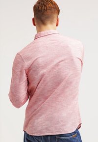 Camicia a maniche lunghe a righe rosse e bianche con una texture morbida, colletto a bottone e orlo leggermente curvo, indossata con jeans blu.