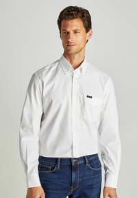 Camicia bianca con bottoni, maniche lunghe, taschino sul petto e un piccolo logo blu navy sulla tasca. Indossata con jeans in denim scuro.
