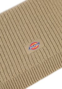 Bonnet en tricot beige avec une texture côtelée. S'agence d'une étiquette en tissu rectangulaire avec le logo Dickies en rouge, jaune et bleu.
