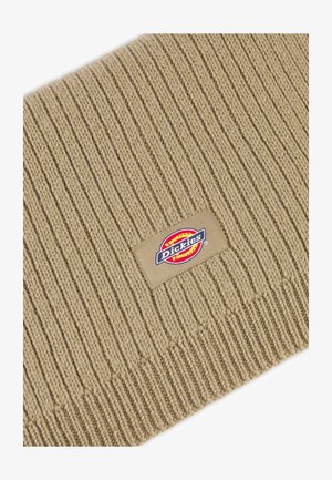 Bonnet en tricot beige avec une texture côtelée. S'agence d'une étiquette en tissu rectangulaire avec le logo Dickies en rouge, jaune et bleu.