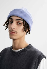 Lichtpaars geribbeld mutsje gedragen met een donkergrijze mouwloze trui over een wit shirt. Kenmerkt zich door zichtbare gedraaide haarstrengen.