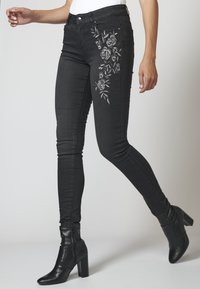 Kvinna som bär svarta skinny jeans med broderad blommönster på låret och svarta klackiga ankelstövlar, som går mot en ljus bakgrund.