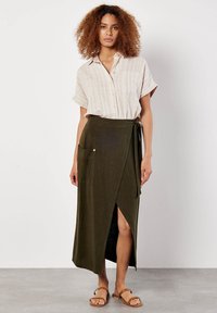 Frau trägt ein beiges, kurzärmliges, geknöpftes Hemd, das in einen olivgrünen Wickelrock mit Taschen gesteckt ist, und braune Sandalen. Sie steht auf einem grauen Boden.